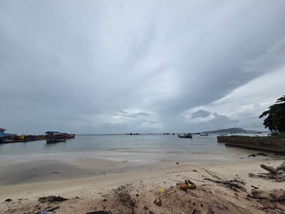 The image depicts a cloudy day scene at a beach in Koh Toch Village, Cambodia. In the foreground, a sandy beach is littered with debris, including plastic, wood, and a broken coconut. The sand shows signs of tidal patterns. The water is calm, with several boats anchored or in motion further out. Some of these boats are larger, with visible structures and multiple decks, while others are smaller, open vessels. In the distance, a shoreline with vegetation and a stone wall is visible. A hilly landmass or island can be seen on the horizon. The sky is overcast with heavy, grey clouds, suggesting a potentially impending rain. The overall impression is a somewhat unkempt beach area in a coastal village.