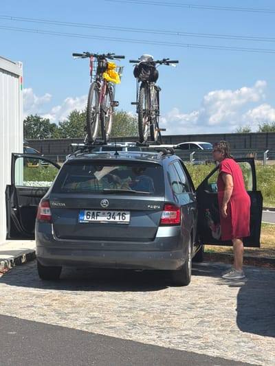An outdoor, eye-level medium shot shows a dark gray Skoda Fabia station wagon parked on a cobblestone surface. The car's rear is visible, with the driver's side door open. A woman wearing a long, red dress and gray sneakers stands beside the open car door, facing away from the camera. Two bicycles are mounted on a roof rack atop the station wagon. The sky is blue with scattered white clouds. In the background, a white barrier and trees are visible. A license plate on the rear of the car reads "6AF 3416". The image is captured during daylight. The exact location is not identifiable within the city of Gralla, Austria, based on the visual information provided.