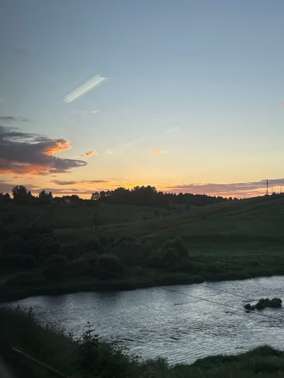 A serene evening scene unfolds over the Volga River near Zubtsov, Russia. The sky transitions from a pale blue at the top to hues of soft orange and pink near the horizon, indicating a sunset. A peculiar, elongated cloud formation, resembling a stretched arrow or a light beam, is visible high in the sky.

The landscape features rolling green hills dotted with trees, creating a natural, undulating terrain. A dark band of trees forms a dense line across the middle ground, separating the hills from the sky. In the distance, some structures, possibly houses or farm buildings, are faintly visible amongst the trees. The foreground is dominated by the tranquil waters of the Volga River, its surface shimmering with the fading light and reflecting the muted colors of the sky. The riverbanks are lined with dark greenery, with tall grass and foliage obscuring the immediate edge. Electrical wires are strung across the river and its banks, connecting to utility poles on the right side of the frame, suggesting the presence of nearby infrastructure. No people or animals are discernible in the image. The overall impression is one of quiet, rural beauty at dusk.