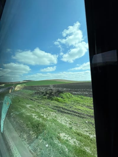 The image shows a view from inside a vehicle, likely a car or truck, looking out through the windshield. The sky is blue with scattered white clouds. The landscape features a mix of terrain including cultivated fields (some with green crops, others recently plowed and dark brown), and grassy areas. The vehicle's interior is visible at the top and bottom of the frame. The location is unspecified beyond a view of outdoor terrain and a sky, implying a location near Karaisen, Bulgaria. No people are visible.