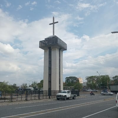 The image displays the Obelisco de Barquisimeto, a prominent monument located on Avenida Libertador in Barquisimeto, Venezuela. The Obelisco is a tall, octagonal concrete tower with a multi-level, glass-enclosed observation deck at its apex, surmounted by a large metal cross. Vertical dark panels or windows punctuate the tower's light-colored concrete facade.

In the immediate foreground, a multi-lane asphalt road stretches horizontally, marked with white dividing lines. A white Chevrolet Silverado-style pickup truck is parked adjacent to the road, near the monument's perimeter. Further along the road, a red motorcycle with a visible rider is in motion, followed by a light-colored sedan and a darker sedan. The front of a white van is partially visible on the far right.

The base of the Obelisco is enclosed by a black metal fence featuring a geometric pattern. Beyond this fence, green trees and foliage are present. In the background, under a partly cloudy sky, additional trees and distant urban structures, including multi-story buildings, are visible. Overhead utility lines are present in the upper right quadrant of the image, alongside a street light fixture. The scene is illuminated by daylight.
