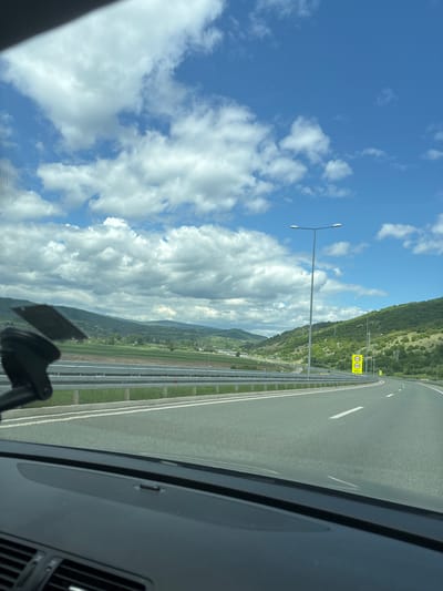 The image shows a view from inside a vehicle, likely a car, traveling on a highway. The sky is predominantly blue with scattered, puffy white clouds. To the right of the sky, there is a green, mountainous landscape. The highway has multiple lanes and a metal guardrail separating the road from the adjacent terrain. Road signs with numbers are visible along the side of the road. The interior of the vehicle is partially visible, showing the dashboard and windshield. The location is near Dimitrovgrad Municipality, Serbia.