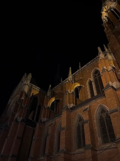 The image shows the co-cathedral of St. Peter and Paul in Osijek, Croatia at night, viewed from a low angle. The co-cathedral is constructed of red brick and features Gothic architectural elements, including pointed arches, intricate stonework, and stained glass windows. The building is illuminated by artificial lighting. The rest of the image is black, suggesting a dark sky.