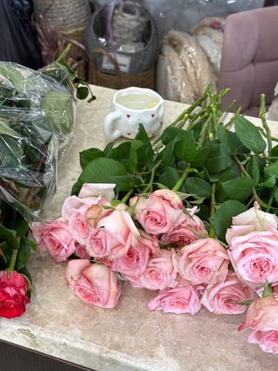 This media file displays a close-up view of a bouquet of light pink roses, arranged on a textured, light brown surface that appears to be a table. The roses vary in their stage of bloom, with some tightly furled and others more open, showcasing their delicate petals. Green leaves and stems are interspersed throughout the bouquet.

In the lower left corner of the image, a portion of another bouquet of flowers, possibly red roses, is visible, partially wrapped in clear plastic. To the left of the pink roses, a small white ceramic mug with red heart accents is positioned, containing a light-colored liquid, likely water. Behind the mug and to the left, other objects are discernible, including what appears to be a basket or container wrapped in burlap or rope, and further back, a blurred pink upholstered object, possibly a chair or couch. The lighting suggests an indoor setting, possibly during the daytime given the brightness. There is no visible text in the image. The scene evokes a sense of freshness and floral beauty, potentially a gift or preparation for an event. The presence of the mug with water suggests the flowers are being kept fresh.