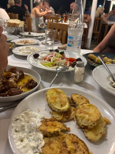 A close-up, eye-level shot captures a table laden with Mediterranean dishes at a restaurant in Ierissos, Greece. In the foreground, a white plate holds several pieces of fried zucchini, sprinkled with coarse salt. To the left, another white dish contains a creamy white dip, possibly tzatziki, with visible green herbs. A larger white plate in the mid-ground displays a Greek salad with tomatoes, cucumbers, olives, and feta cheese, along with a silver salt shaker. To the left of the salad, a white bowl holds a portion of meat and mushroom stew. In the background, out of focus, several people are seated at tables, some of them eating and interacting. The overall ambiance suggests a lively evening meal at a taverna.