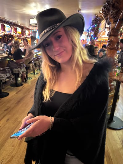 A fair-skinned woman with blonde hair and a black cowboy hat gazes at the viewer, holding a cell phone. She is wearing a black top and a dark cardigan or shawl with fur trim. Behind her, a bar is visible, populated with patrons and decorated with lights. The scene suggests a festive or nighttime setting.