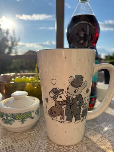 The image shows a close-up view of a white ceramic mug with a printed illustration of mice dressed in formal attire. The mug is adorned with small grey dots. To the left of the mug is a small white sugar bowl with green and white patterned trim. In the background, a glass bowl filled with green grapes is visible, and behind that, a large bottle of dark soda is positioned. The scene appears to be set on a table covered with a white lace tablecloth, outdoors. The background suggests a sunny day with a bright sky and trees, possibly indicating a balcony or patio setting. The specific location in Novaya Usman, Russia is not depicted or implied in the visual elements of the image.