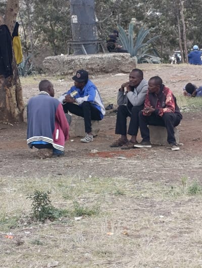 An outdoor scene in Matungulu west ward, Kenya, depicts six individuals in varying states of gathering and rest on dry, patchy ground with sparse vegetation.
In the foreground to midground, four men are clustered. One male, wearing a blue and maroon long-sleeved shirt and blue shoes, is squatting on a small stone with his back partially to the viewer, facing the others. Opposite him, a male wearing a blue and white athletic jacket, black pants, a black baseball cap, and grey athletic shoes, sits on a rectangular grey stone block, holding a metallic can in both hands. To his right, another male in a grey hooded sweatshirt, black pants, and dark sandals, sits on a similar stone block with his hands clasped under his chin. Adjacent to him, a fourth male wearing a dark red, black, and white patterned long-sleeved shirt, black pants, and dark low-top shoes, also sits on a grey stone block with his hands clasped. These four individuals appear to be engaged in an interaction or discussion.
In the mid-background, to the left, a dark jacket and a yellow garment hang from a tree trunk. Behind the main group, a dark, cylindrical metal structure, possibly a utility component, is visible atop a large, irregular stone base, partially obscured by trees. Large agave-like plants are present in the background. Further back, two additional individuals are visible: one wearing blue clothing, partially obscured, appears to be resting on the ground to the far right, and another individual in dark clothing is lying on a larger stone structure behind the main group. The ground consists of dry grass, bare earth, and scattered small debris. The sky is overcast.
