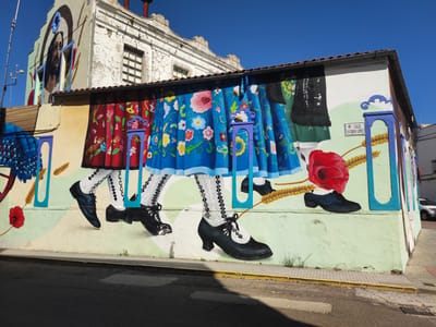 This media file features a vibrant mural painted on the side of a building in Valencia de Alcántara, Spain. The mural depicts a portion of a traditional folk dance, focusing on the colorful skirts and intricately patterned stockings and shoes of dancers. The skirts are vividly colored with floral motifs, and the stockings feature geometric and lace-like designs. The dancers are wearing black, heeled shoes.
The mural is set against a backdrop of painted elements that suggest a stage or an outdoor setting, with stylized representations of wheat stalks and a bright red poppy. Above the mural, the building itself is weathered, with peeling paint and visible cracks, contrasting with the lively artwork below. On the upper left section of the building, a painting of a human face, possibly religious in nature, is also visible.
A street sign with the text "CALLE ESTEBAN LOPEZ" is painted onto the mural, indicating the location. The scene is bathed in bright sunlight, casting shadows on the asphalt road in the foreground, suggesting it is daytime. The sky is a clear, bright blue, indicating fair weather. The overall impression is one of cultural celebration and artistic expression in a Spanish town.