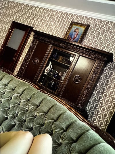 The image shows an ornate, dark wood cabinet with glass doors displaying various decorative items, including a white pitcher with red and blue details. The cabinet is set against a patterned wallpaper and next to a tufted, green velvet armchair. A framed painting of a figure in flowing fabric, possibly a mythological scene, hangs on the wallpaper to the left of the cabinet. The lighting suggests an indoor setting, possibly a home or a formal room. The overall impression is one of antique or vintage decor. The composition is angled, and a portion of a person's legs and knees are visible in the lower right corner, resting on the armchair. The image is taken from a high-angle perspective, looking down at the scene.