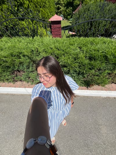This is a vertical, medium shot selfie taken outdoors on a sunny day in Chahor, Ukraine. The subject is a young woman with long, dark hair, wearing glasses and a blue and white striped dress. She is looking at the camera with a slight smile. The setting appears to be a park or garden, with lush green bushes on the left and a paved path on the right. A black wrought-iron fence with decorative swirls runs along the edge of the bushes, and a small, red-roofed structure, possibly a birdhouse or decorative element, is visible behind the fence. The woman is wearing sandals and a bracelet with various beads and charms. The lighting suggests it is daytime, likely late morning or afternoon, given the strong shadows.