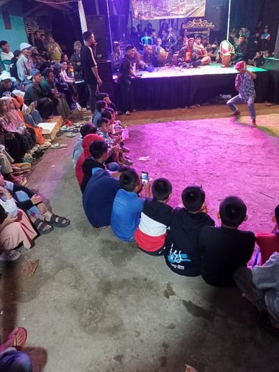 This image captures a vibrant nighttime cultural performance in Kecamatan Cimaung, Indonesia. A large audience, comprising both adults and children, is gathered to watch a traditional music and dance or martial arts display.

The scene is primarily illuminated by artificial light, with a striking magenta/purple hue cast across the dirt ground directly in front of the stage. On a raised stage, several male performers are actively engaged. Some are playing traditional musical instruments, notably various drums, while another man stands near the stage's edge, possibly acting as an emcee or director. In front of the stage, a performer dressed in patterned clothing and a red head covering is captured in a dynamic, low stance, indicating a dance or martial arts sequence.

The audience is seated on the ground and on low benches or platforms surrounding the performance area. Many children occupy the front rows, sitting directly on the ground and watching with keen interest. Some children are visible holding mobile phones, capturing the event. Adults are seated further back, also appearing engaged and attentive.

The time of day is clearly night, evidenced by the reliance on artificial lighting. The setting appears to be an informal outdoor or semi-outdoor venue, suggesting a community gathering or local festival. In the background, behind the stage, a banner with text is visible, likely in Indonesian. While most of the text is illegible due to distance and lighting, it appears to feature words related to art and culture. The overall atmosphere is lively and communal, centered around a traditional artistic display.