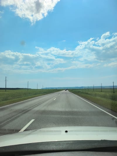 The image depicts a view from a vehicle's perspective looking down a paved, two-lane road that stretches into the distance under a bright blue sky with scattered white clouds. The road is flanked by flat, green fields, possibly farmland, with rows of utility poles lining the right side of the road. In the distance, the landscape appears to be rolling hills. A few vehicles are visible on the road ahead. The foreground shows the dashboard and hood of the vehicle from which the photograph was taken, with a portion of the windshield visible. There are no people clearly visible in the image. The scene is consistent with a rural or semi-rural environment, potentially along a highway outside of a populated area. It is not possible to determine the specific location in the city of Покровка, Russia, from the image alone, as there are no identifying landmarks or signs.