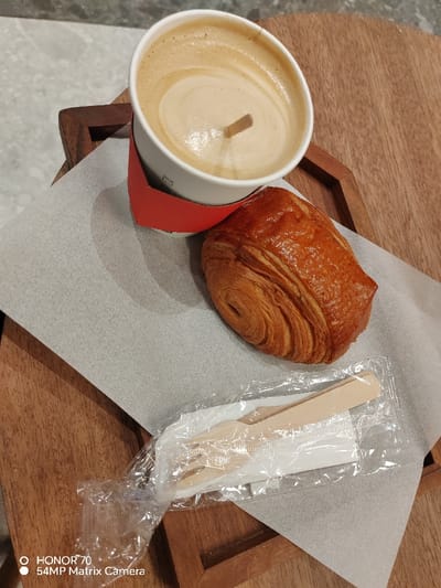 A paper cup of coffee with a red sleeve sits alongside a golden-brown croissant on a square wooden tray lined with grey parchment paper. A wrapped wooden fork and knife set rests at the bottom of the tray. The coffee shows a swirl of light brown color on top and appears to have a wooden stick in it. The tray has a raised edge and sits on a grey surface in the background.