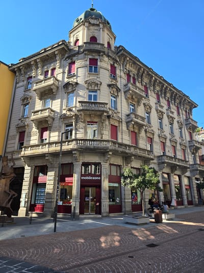 This media file captures a sun-drenched street scene in Bellinzona, Switzerland. The main subject is an ornate, multi-story building with a distinct architectural style, featuring intricate stonework, decorative balconies, and numerous windows with red shutters. A green domed structure is visible on the left side of the building's facade. The building appears to house several businesses on its ground floor, including "mobilezone" and "wingo".
In the foreground, a cobblestone street is illuminated by bright sunlight, casting sharp shadows. Two people are visible on a bench near the building: one is seated, and the other is partially obscured. Another person is on a bicycle in the middle ground. A small tree is growing in front of the building, adding a touch of nature to the urban environment.
The time of day appears to be late morning or early afternoon, given the angle and intensity of the sunlight. The sky is a clear, vibrant blue, indicating fair weather. The overall atmosphere is one of a typical day in a European town, with everyday activities taking place against a backdrop of historic architecture. No strong emotions are conveyed, but the scene suggests a peaceful and functional urban setting.