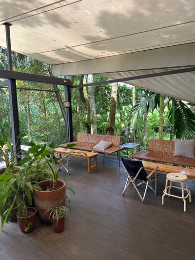 The image shows an outdoor living space under a covered structure, possibly a patio or veranda, located in Licin, Indonesia. The space is furnished with woven benches with cushions, a small folding camping chair, and a white stool. Several potted plants of varying sizes, including ferns, are placed around the seating area. The structure has a dark metal frame. Beyond the structure, a lush green environment with numerous trees and plants is visible. The floor is made of dark-stained wooden planks. There are no people visible in the image.