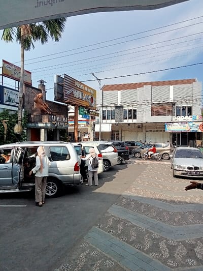 The image shows a street scene in Kudus, Indonesia, located at coordinates -6.802 latitude and 110.844 longitude. The scene is captured from the perspective of a pedestrian standing on the sidewalk, looking across the street. There are multiple parked cars on both sides of the street. A silver SUV with its rear passenger door open is parked closest to the viewer. Two women, wearing headscarves, are standing near the SUV. The woman closer to the viewer is facing the camera and the woman further away is facing the SUV. There is a man with a helmet on his head standing on the other side of the street, near a white motorcycle. He is facing the parked cars. In the background, a building with a sign that reads "PISA JUMBO MIRAJ" in Indonesian, is visible. Several other buildings can be seen in the distance. The street is paved with patterned gray stones. There are power lines running across the sky. The sky is clear and blue. The scene shows a typical street in Kudus, Indonesia with a mix of vehicles and pedestrians.