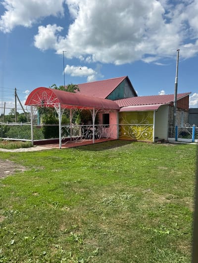 The image depicts a rural scene in Yazykova Pyatina, Russia, on a sunny day with scattered clouds. The foreground is a well-maintained lawn with green grass. Behind the lawn, a house with a red tiled roof is visible. The house has a light-colored exterior with green and yellow accents. A red-covered porch with white decorative railings extends from the house, providing a sheltered outdoor seating area. Next to the porch, there is a section of the house with yellow siding and decorative white fencing. To the right of the house, a gray metal fence and a smaller white and blue gate are present. The background shows trees and power lines, typical of a village setting. There are no people visible in the image.
