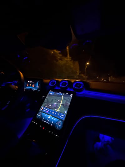 The image captures the interior of a car at night, focusing on the dashboard and infotainment system. The primary subjects are the illuminated car interior elements, including the steering wheel, air vents, digital instrument cluster, and a large central infotainment screen.

The scene is set inside a modern vehicle, characterized by prominent blue ambient lighting tracing the dashboard lines and illuminating the circular air vents. Outside the car, through the windshield, streetlights are visible, suggesting a nighttime setting. The ground outside appears light-colored and somewhat blurry, possibly indicating snow, frost, or environmental conditions like fog.

An activity of driving or being parked at night is taking place. The infotainment screen displays a navigation map, suggesting travel, and music is actively playing, with "Ace of Base" and "Ace of Base - Ha..." visible as the artist and song title. The climate control is set to 21.0 degrees Celsius and shows "АЦТО" (Auto) and "Меnu климат" (Menu climate). The time displayed on the screen is 22:57. The digital instrument cluster, partially visible behind the steering wheel, shows a "29" and a "3," likely related to vehicle speed or gear selection. Additional text on the infotainment screen includes "Навигация" (Navigation), "Телефон" (Phone), and "iPhone на". The location context provided is Zvezditsa, Bulgaria, aligning with the Cyrillic text visible on the screens.
