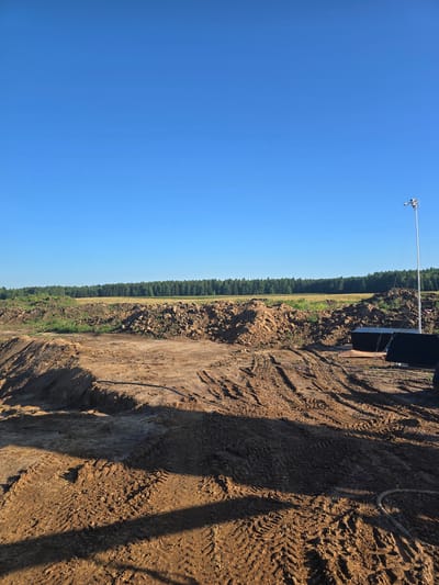 This image depicts an outdoor construction site under a clear blue sky. The foreground is dominated by disturbed earth and tire tracks, indicating recent activity by heavy machinery. In the mid-ground, there are piles of dirt and some green vegetation. Beyond that, a line of trees forms the horizon. To the right of the image, a tall pole with a camera and some dark, rectangular structures are visible, suggesting temporary site security or monitoring equipment. The location is identified as Piecnik, Poland. There are no people visible in the image.