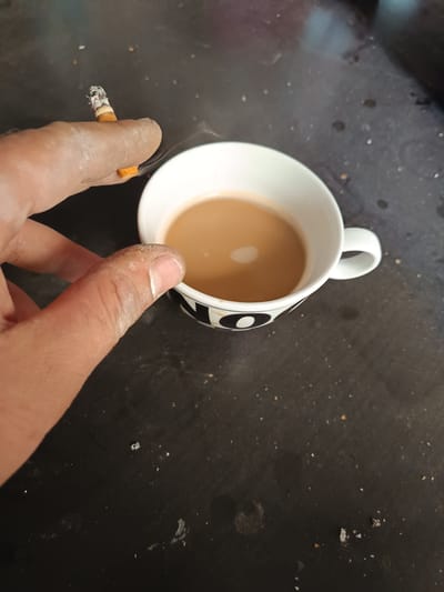 A close-up shot captures a person's hand holding a lit cigarette over a white mug filled with a light brown beverage, likely coffee or tea. The mug has a black and white patterned rim. The background is a dark, speckled surface, possibly a table. The scene appears to be indoors, with no specific geographical markers to indicate a location in Frechen, Germany. The lighting is direct, highlighting the ash on the cigarette and the surface of the liquid. The hand shows signs of a powdery substance, possibly dust or chalk.