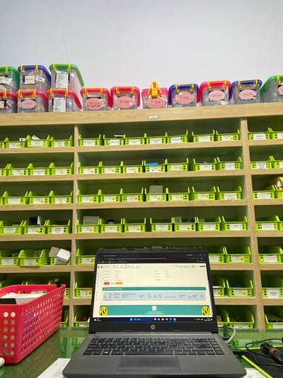 This is a forensic, matter-of-fact description of a room that appears to be a pharmacy or medical storage area, likely located in Margoyoso, Indonesia. The scene is orderly and well-organized, suggesting a professional setting. **Environment:** The room is brightly lit with white walls and wooden shelves lining the walls. The shelves are filled with numerous green plastic bins and some larger clear plastic containers with colored lids. The bins are neatly arranged, each containing various medical supplies or medications, many with labels indicating their contents. A desk is positioned in the foreground, on which a laptop computer is open, displaying a medical or pharmacy-related software interface. Cables and a red storage basket are also present on the desk. The flooring appears to be a green, reflective surface, possibly a countertop or desk. **Objects:** * **Shelving:** Multiple rows of wooden shelves are visible, filled with organized containers. * **Containers:** Numerous bright green plastic bins are uniformly placed on the shelves. Larger, clear plastic storage containers with blue, yellow, and pink lids are stacked on the top shelves. Many of these containers have handwritten labels or printed slips of paper attached. * **Medications/Supplies:** The bins contain items that appear to be pharmaceuticals or medical supplies, based on the labeling and packaging visible. * **Laptop:** A black HP laptop is open on the desk, displaying a software interface with fields like "Nama Pasien," "PMR," and tables with columns such as "Tgl Resep," "Nama Resep," and "Jenis Kamar." * **Cables:** Black and green cables are present near the laptop, suggesting electronic devices are connected. * **Red Basket:** A bright red, perforated plastic basket is on the desk, containing white boxes or packaging. * **Labels:** Numerous labels, some handwritten and some printed, are affixed to the containers, indicating the contents and possibly classifications of the stored items. Examples include "SPINAL," "10. GA," "7. GA," "ACARBOSE," "METFORMIN," "AMOXICILLIN," "PARACETAMOL," and specific dosages. **Actions/Interactions:** The scene is static, implying no immediate human activity. The laptop is open and displaying information, suggesting it is in use or has recently been used for managing inventory or patient records. The organization of the shelves and bins indicates systematic storage and retrieval of medical supplies. **Location Context (Margoyoso, Indonesia):** The presence of Indonesian language text on the laptop screen, specifically terms like "Nama Pasien" (Patient Name), "PMR" (likely Patient Medical Record), and "Resep" (Prescription), strongly indicates the location is Indonesia. Given