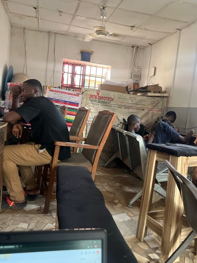 **Scene Description:**
The image depicts a small, cluttered room in what appears to be a computer or internet cafe. The room has tiled floors and pale walls. A promotional poster advertising "UNIQUE CHIPSETS INFOTECH STARLINK SERVICES" and other computer-related services is prominently displayed. The poster lists data subscription prices, computer training, graphic design, and internet services.
Several young men are present. One man is leaning on a table, seemingly asleep. Another two men are sitting in chairs, one with his feet up and seemingly looking at a device. Additional individuals are possibly seated.
The location is possibly in Barako, Nigeria. The poster mentions "FUT Minna, Bosso Niger State," which suggests a connection to the Federal University of Technology, Minna, located in Bosso, Niger State, Nigeria. Bosso is near Barako. The poster also has contact information and a list of services that are provided. The chairs are a mix of wooden and metal-framed. A computer screen is partially visible in the foreground.
