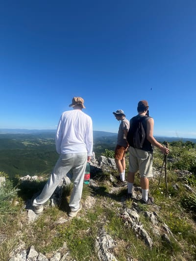 Three individuals are standing on a rocky, grassy precipice overlooking a valley with rolling, forested hills under a clear blue sky. The person on the left, viewed from the back, wears a tan bucket hat, a long-sleeved white shirt, and grey sweatpants. A green plastic bottle of water is held in their right hand. In the middle, another individual, facing left and slightly away from the camera, wears a grey bucket hat, a grey t-shirt, and orange shorts. The person on the right, facing away from the camera and holding two wooden hiking poles, wears a brown baseball cap, a dark tank top over a grey t-shirt, and light beige shorts. They are holding a mobile phone in their right hand, appearing to take a photo or video. The environment is natural and mountainous, with sparse vegetation on the rocky terrain. The scene suggests a hiking or outdoor exploration activity.