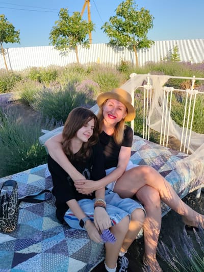 A woman and a teenage girl are sitting on a blanket spread on the ground, with a white metal bed frame behind them. The woman, wearing a straw hat, a black t-shirt, and white shorts, has her arm around the girl. The girl, with her eyes closed and a slight smile, is wearing a black t-shirt and blue denim shorts. They are in a field of lavender, with trees and a white fence in the background. The sun is setting, casting long shadows and creating a warm, golden light. The overall mood is one of affection and relaxation, possibly during a summer evening in Nechaiane, Ukraine. A leopard-print bag and bracelets on the girl's wrist are visible.