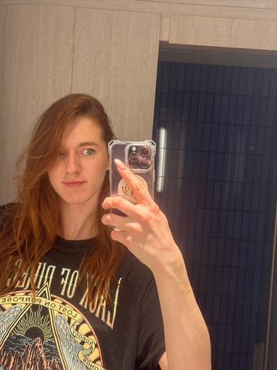 A woman takes a mirror selfie in a bathroom. She holds her phone up with her right hand, partially obscuring her face. She has red hair and a black t-shirt with a graphic design. The background consists of a wall on the left and a dark, reflective surface above, possibly a window or panel. The lighting appears to be bright and possibly artificial.