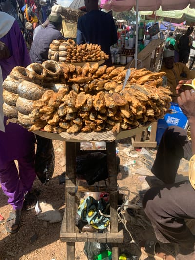 This is a horizontal, eye-level photograph taken outdoors in what appears to be a market setting. The foreground is dominated by a large platter filled with fried fish. The fish are small, whole, and covered in a golden-brown batter, some piled high and others arranged in a circular fashion on the platter. A silver serving spoon is stuck into the fish.
Behind the platter of fish, a wooden stand holds more items, including what looks like more fried fish, bundles of dried fish, and bags of merchandise. The ground around the stand is dusty and littered with debris.
Several people are visible in the background, dressed in various types of clothing. One person's hands are visible in the upper part of the frame, holding a slice of lime and wearing a silver ring. Another person, wearing a dark blue top and a head covering, is partially visible to the left. Further back, several other figures are present, some in more colorful attire. The scene suggests a bustling market atmosphere, likely during daylight hours, given the strong shadows.
The overall impression is a vibrant, active marketplace in Nigeria, focusing on the sale of food, specifically fried fish, and other goods. There is no visible text in the image.