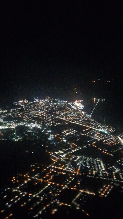 This image captures an aerial perspective of a city at night, presenting a sprawling network of artificial lights against a predominantly dark backdrop. The urban area occupies the right and bottom portions of the frame, with the upper-left quadrant remaining in deep shadow, likely representing the night sky or unlit natural landscapes.
The city lights, predominantly glowing in warm yellow-orange and bright white, form intricate patterns that delineate streets, blocks of buildings, and infrastructure. These luminous lines and clusters illustrate the layout of the city, suggesting a grid-like or organic development. A notable detail is a distinct linear stretch of lights extending from the main urban mass into the dark expanse, possibly indicating a long pier, a bridge, or a coastal feature along the water's edge, aligning with Port Dickson's coastal location in Malaysia. Scattered individual lights are also visible further out in the dark, hinting at isolated structures or possibly boats.
The image conveys the vastness and organized complexity of an urban environment seen from above during nighttime. The visibility is good, suggesting clear weather conditions. No specific human activity or identifiable objects beyond the city lights are visible, nor is any text present. The overall impression is one of nocturnal urban expansion and connectivity.