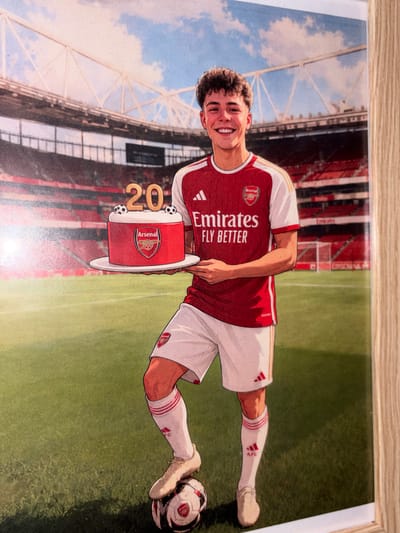 The image shows a framed cartoon drawing of a soccer player wearing an Arsenal jersey, white shorts, and matching socks and shoes. He is holding a cake that resembles a drum with the Arsenal emblem on it, adorned with the number "20". A soccer ball is on the ground next to his right foot. The background appears to be a soccer stadium with a cloudy sky above it. There is no information in the image indicating a location in Bezden, Bulgaria.