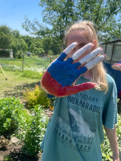 A young woman with blonde hair pulled into a ponytail holds her right hand up in front of her face, obscuring her features. Her hand is painted with the colors of the Russian flag: red on the palm and thumb, blue across the fingers, and white on the tips of her fingers. She is wearing a teal t-shirt with white text that reads "LEGENDARY SUMMER PARADISE" and partially obscured text below. She is outdoors in a garden setting with lush green foliage and trees. The sky is bright blue and clear. In the background, there are small wooden structures, possibly sheds or outbuildings, and a greenhouse. The scene appears to be set on a sunny day.
