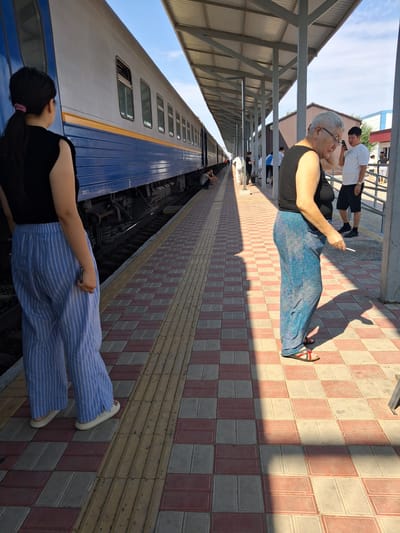 At a train station platform in Maqat, Kazakhstan, a young woman with dark hair pulled back in a ponytail stands in the foreground, wearing a black sleeveless top and light blue and white striped wide-leg pants. She faces away from the camera, looking towards a parked train. To her right, an older woman with short gray hair and glasses wears a black tank top and blue patterned wide-leg pants. She holds a white cigarette in her right hand and looks down, seemingly at her phone, which is in her left hand. Behind her, a young man in a white t-shirt and black shorts stands, also looking at his phone. The platform is paved with alternating red and light-colored rectangular tiles, with a textured yellow tactile paving strip along the edge. A covered walkway with metal supports runs along the platform, casting long shadows. The train, with blue and yellow detailing, is visible on the tracks. Other people are visible in the background, some sitting on the platform and others walking. The sky is clear and blue.