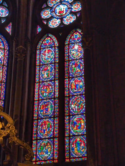 The image shows a view of stained glass windows inside a church, likely a cathedral, given their scale and intricacy. The windows are tall and narrow, divided into several circular panels, each depicting a scene with figures. The colors are vibrant and varied, dominated by blues, reds, and yellows, with some green and white.
The main focus of the image is two large, vertical stained glass windows. The left window is partially obscured by a large, ornate golden architectural element on the left side of the frame, which appears to be a balustrade or choir screen. The right window is fully visible. Both windows feature a repeating pattern of circular medallions containing narrative scenes. These scenes likely depict biblical stories or lives of saints, a common theme in church stained glass. The architectural style of the church appears to be Gothic, suggested by the pointed arches of the windows and the dark, imposing stonework visible in the background.
The image is taken from a low angle, looking upwards towards the windows. The lighting is dim, with the stained glass illuminated by an unseen light source, possibly sunlight filtering through. This creates a dramatic contrast between the illuminated glass and the dark interior of the church. The overall impression is one of solemnity and grandeur.
The question about Urvillers, France is not addressed by the visual information present in the image. There is no contextual information within the image itself that would indicate its specific location in Urvillers or any other city.