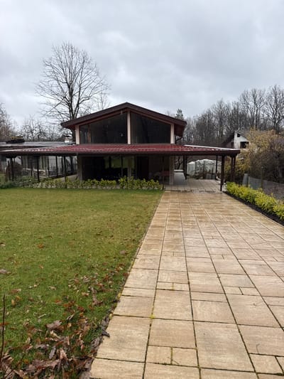 An exterior daytime shot in Peyovstsi, Bulgaria, depicts a two-story modern house under an overcast sky with visible light precipitation. The house features a dark red tiled roof, dark brown eaves, and extensive dark-tinted glass windows on both levels. A dark wooden pergola extends from the right facade, sheltering a portion of a wet, light-colored paver driveway composed of rectangular and square blocks, which leads towards the structure. To the left of the driveway is a vibrant green lawn with scattered brown leaves. A low, manicured green hedge lines the base of the house, and another row of yellow-green shrubs runs parallel to the driveway on the right, adjacent to a wire fence. Bare deciduous trees form a dense backdrop, with a partially visible, older white house on the far right among the trees. No individuals are present.