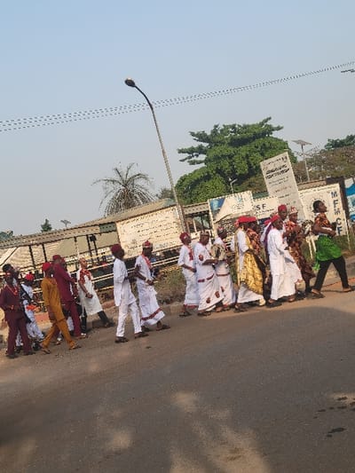 The image depicts an outdoor daytime scene in San Martín, Colombia, showing a procession of approximately 15-20 individuals walking from left to right along an asphalt road. Most individuals are adults dressed in traditional or ceremonial attire, featuring white robes, some with red trim or sashes, and several wear red caps or hats. Distinct outfits include a dark red suit, a mustard-yellow tunic and trousers, a golden-yellow patterned garment worn over white, and a green patterned top. Some individuals appear to be carrying small objects, possibly musical instruments.
The road surface in the foreground is asphalt. To the right of the procession, there is a raised concrete curb and a narrow strip of dirt and sparse vegetation. In the mid-ground, a tall, dark gray street light pole curves upwards towards the center. Behind the procession, various structures are visible, including what appears to be a building with a corrugated roof on the left. Several white signs and billboards are present, some partially legible with letters like "SF" and "MFM." The background features dense green foliage from trees, including a prominent large tree and a palm tree. Overhead, multiple thin, dark lines, likely electrical wires, traverse a clear, light blue sky. The scene is illuminated by bright daylight.