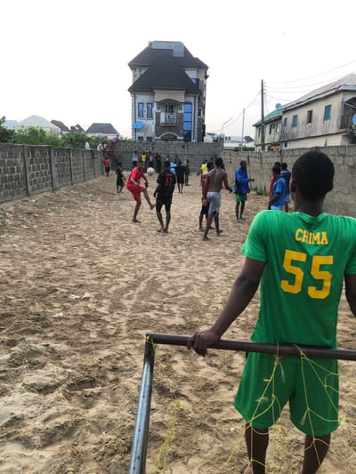 A group of people are playing beach soccer on a sandy beach. The player closest to the viewer is wearing a green jersey with the name "CHIMA" and the number "55" on the back. Another player is kicking the ball. A net is in the foreground, with a person standing behind it. Buildings and a wall can be seen in the background.