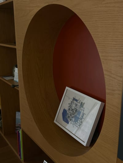 The image shows an overhead view of a wooden coffee table with an oval-shaped depression featuring a red inner surface. Resting partially on the red surface and partially on the surrounding wood is a framed picture with a white border, depicting what appears to be a schematic drawing with blue details. The table is part of a shelving unit, with books and other items visible on the shelves below. The location in Mazatlán, Mexico, is not discernible from the image alone.