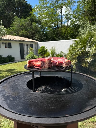 A cut of beef rests on a metal grate inside a grill. The grill is positioned outdoors on a grassy lawn, with trees, bushes, and a two-story house visible in the background. The house is white with a tiled roof. The scene is located in La Couarde-sur-Mer, France.