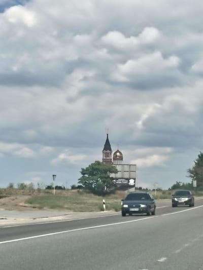The image shows a cloudy sky over a highway and a church in Velykyi Dalnyk, Ukraine. Two cars are driving on the highway, one in the left lane and one in the right lane. In the background, there is a church with a golden dome and a steeple, as well as some trees and bushes. There is also a billboard with some text on it. The overall scene is one of a rural or suburban area on a cloudy day.