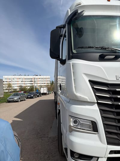 The image shows the front of a white truck parked on a street lined with parked cars. In the background, there is a tall apartment building with multiple windows. The sky is blue with some clouds. The image appears to be taken in Tavry, Russia, judging from the license plates of the parked cars.