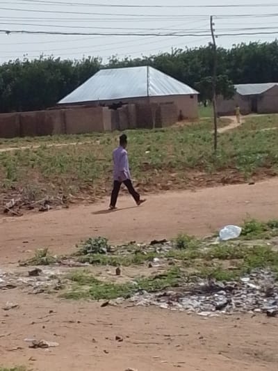 A lone individual walks across a dirt path in a rural setting. They are wearing a light purple long-sleeved shirt and dark trousers. The scene is outdoors with a cloudy sky overhead, suggesting it might be late morning or early afternoon. In the background, there are several simple structures with corrugated metal roofs and brown walls, typical of residential buildings in the area. A line of trees forms a backdrop behind the buildings. The ground is a mix of dirt and patchy vegetation, with some scattered debris, including a white plastic bag visible on the right side of the path. A utility pole with multiple wires stretches upwards on the right side of the image. Another person, smaller in the distance, is also visible near one of the buildings. The overall impression is of a quiet, everyday scene in a Nigerian village.