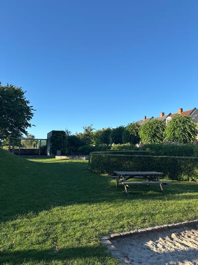 This image depicts a park area on a clear, sunny day. The sky is a vibrant, cloudless blue. In the foreground, there is a patch of sandy ground, bordered by a low brick edge. Beyond the sand, a well-maintained lawn stretches out, with some scattered dry leaves. A weathered wooden picnic table with attached benches is situated on the grass, casting a shadow. In the middle ground, several meticulously trimmed hedges are arranged in layered patterns, forming geometric shapes. Behind the hedges, a concrete structure covered in ivy stands, with a trash receptacle next to it. Further back, a wooden structure with seating and metal fencing can be seen. A large, leafy green tree dominates the left side of the mid-ground. In the background, a row of houses with red-tiled roofs and trees lining the street is visible. The overall impression is of a peaceful, well-kept urban green space. This park area is located in Horsens, Denmark.
