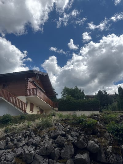 This is a medium-shot, eye-level, outdoor photograph taken on a sunny day with scattered clouds in Les Rousses, France. The image features a wooden chalet-style house with a red-brown exterior, a dark roof, and a balcony with a wooden railing. A white staircase with wooden handrails leads up to the balcony. The house is situated on a rocky slope covered with green vegetation and moss. In the background, there are dense green trees and hedges. The sky above is a vibrant blue with fluffy white and grey clouds. No people are visible in the image.