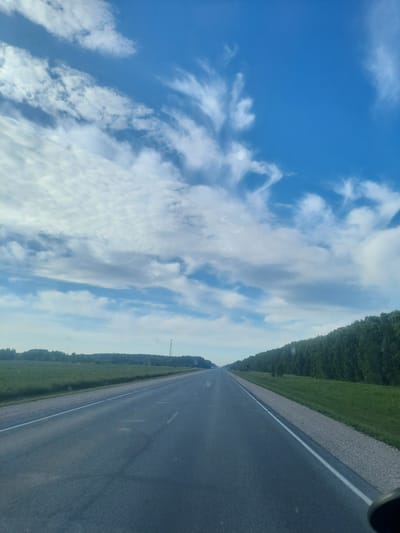 The image depicts a view from a vehicle traveling on a paved road in the city of Pyatiletka, Russia. The road stretches into the distance, bordered on the right by a dense line of green trees and on the left by a grassy field. The sky above is a vibrant blue, partially filled with wispy white clouds. The lighting suggests it is daytime, with the sun likely positioned in the sky, casting subtle shadows. No people or specific structures, apart from a distant tower-like object on the left horizon, are clearly visible in the scene. The road surface shows some wear and tear, with visible patching and lane markings. The overall impression is of a scenic drive through a rural or semi-rural area of Pyatiletka.