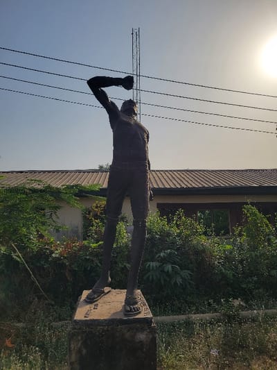 A dark-colored, likely bronze, male statue stands on a two-tiered concrete pedestal under a clear, light blue sky in San Martín, Colombia. The figure faces slightly right, its head tilted upward, with its left arm raised and bent at the elbow, forming a clenched fist. The statue wears sandals and appears bare-chested. In the mid-ground, a building with a reddish-brown tiled roof and light-colored walls is partially obscured by dense green foliage. An open window frame is visible on the building's facade. Multiple dark, horizontal utility lines stretch across the upper frame, and a tall, metal lattice tower is visible vertically behind the statue's upper body. The bright sun is prominent in the upper right quadrant of the image. The ground surrounding the pedestal is covered in dry grass and sparse green vegetation.