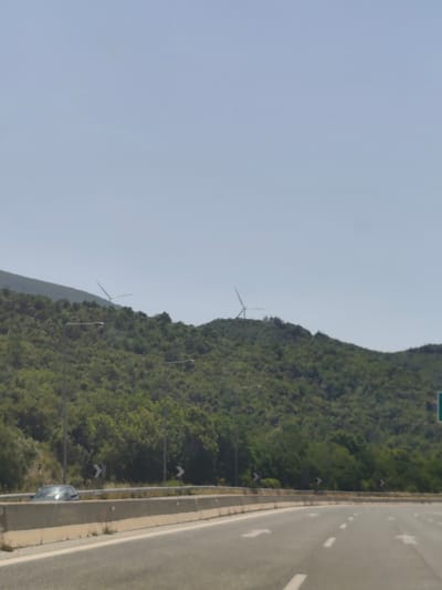A highway scene in Δημοτική Ενότητα Προμαχώνος, Greece. A two-lane asphalt road curves to the right, with white lane markings and directional arrows. A concrete barrier separates the lanes from the shoulder. Beyond the barrier, a silver car is visible. The road is bordered by a low concrete wall and a grassy verge. In the background, a lush green, tree-covered hillside rises steeply. Two wind turbines are visible on the crest of the hill, with their blades extended. Several streetlights are also present along the hillside. The sky is clear and light blue.