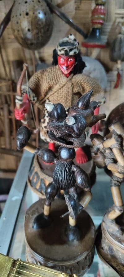 The image displays a close-up of handcrafted figures, likely souvenirs or decorative items, set in a display environment within Cikanyere, Indonesia. In the central foreground, a dark brown, stylized figure is present, constructed primarily from carved coconut shell pieces and lighter wood segments for articulation. This figure possesses an intricate, dragon-like or monstrous head with small, light-colored protrusions and an open mouth. Its body is segmented, suggesting muscle definition, and its arms are raised, with one arm extending forward. The figure stands on a circular, dark brown base.
Behind this figure, a larger, human-like doll is positioned. This doll features a distinct red face with white eyes and mouth markings, a black wig-like hair piece, and a patterned head covering, possibly leopard print. Its torso is covered by a light brown, woven garment, also trimmed with a leopard print. The left hand of this doll holds a small, light brown bow with a red element attached, while its feet are painted red. It stands on its own circular base.
In the upper background, partially visible, is a large, dark, spherical object with multiple small, circular perforations, likely another carved coconut shell. To the upper right, part of a red and gold painted wooden object is discernible. The figures are displayed on a light, reflective surface, possibly glass or polished metal, with a blurred background consisting of vertical and horizontal wooden or bamboo structures.